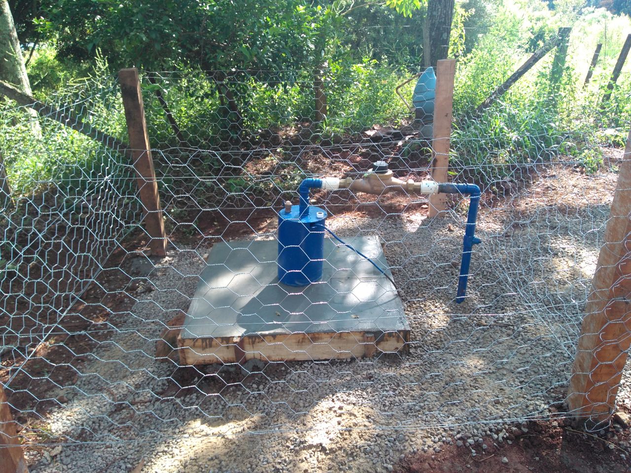 Poço artesiano com cavalete hidráulico azul e hidrômetro instalados sobre base de concreto, cercado por tela de arame em área rural.