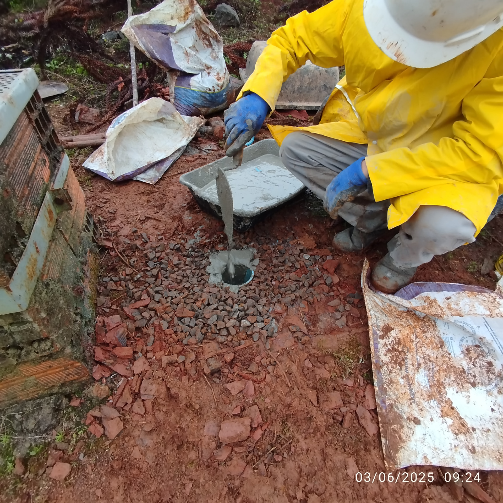 Operário aplicando material de selagem no poço tubular com uso de luva, capacete e avental amarelo de proteção.