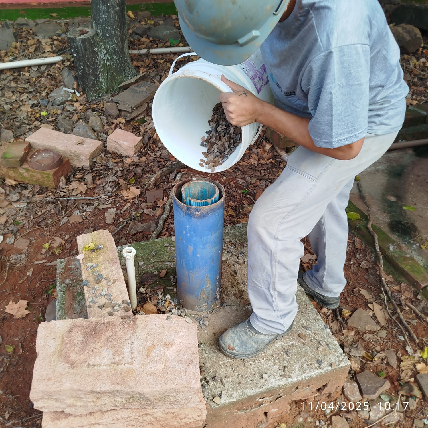 Operário despejando brita e material de preenchimento dentro do revestimento azul do poço tubular.