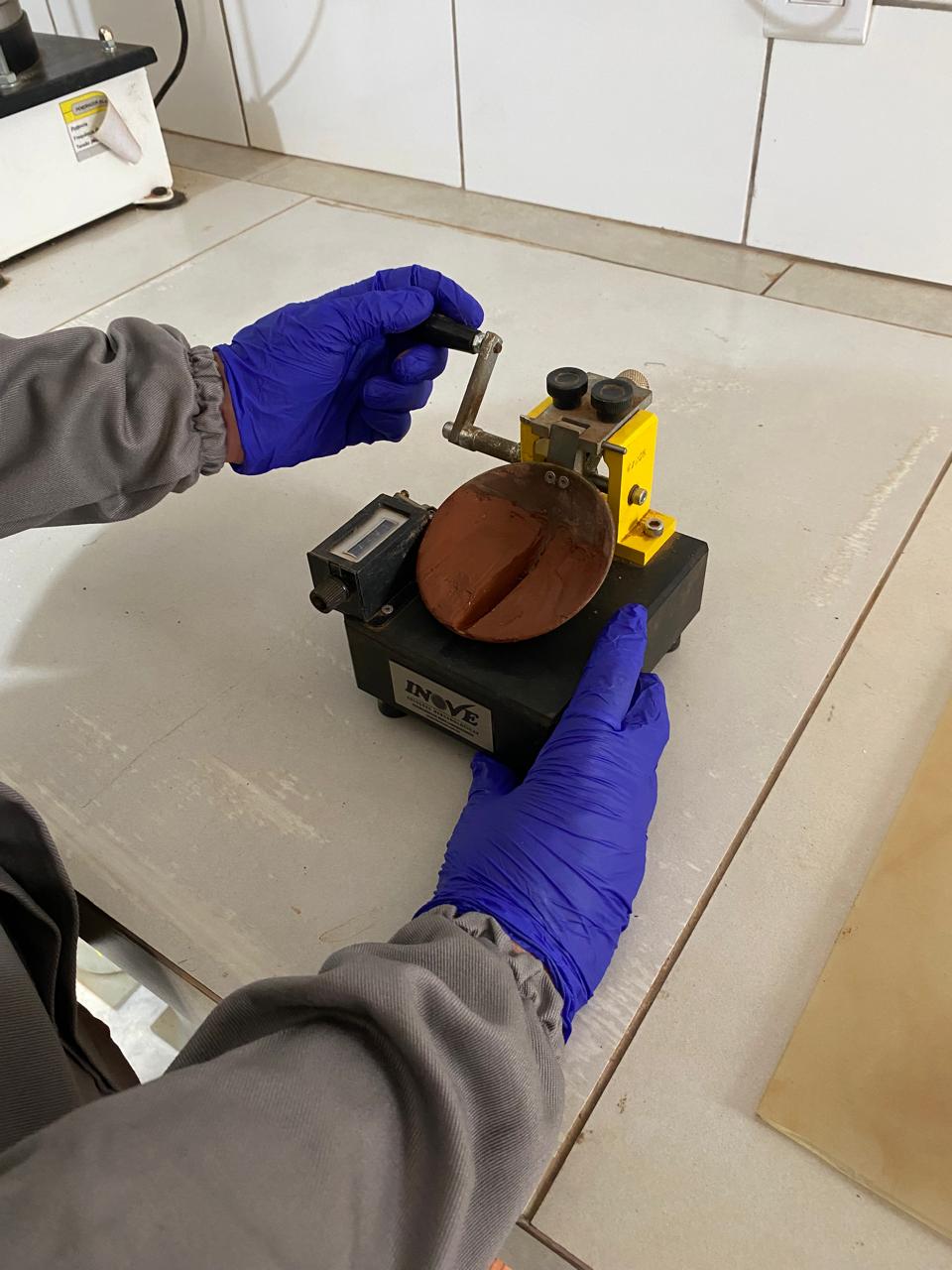 Técnico realiza a determinação do limite de liquidez do solo em aparelho de Casagrande, com amostra de argila em um recipiente metálico de cor cobre sobre base amarela e preta.